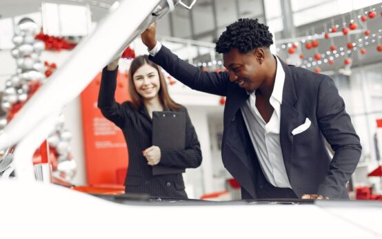 Positive smiling diverse stylish man and female dealer checking under car motor hood and smiling while choosing new car in car showroom
