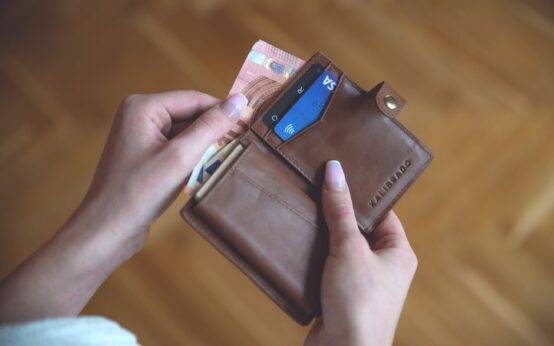 Hands holding a leather wallet with cash and a credit card on a wooden surface.