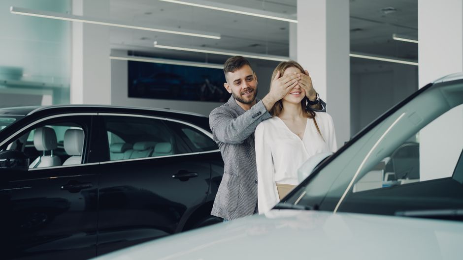A joyful couple enjoying a surprise moment at a car dealership, surrounded by sleek vehicles.